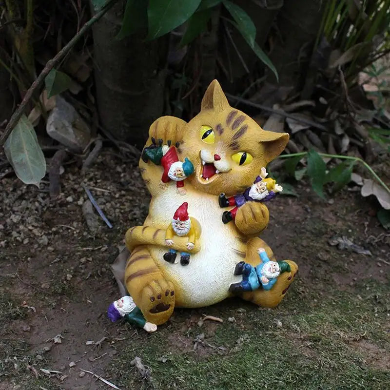 A playful cat statue is surrounded by small gnome figures, positioned in a garden setting. The cat appears cheeky, with a wide smile, as it holds and interacts with the gnomes.