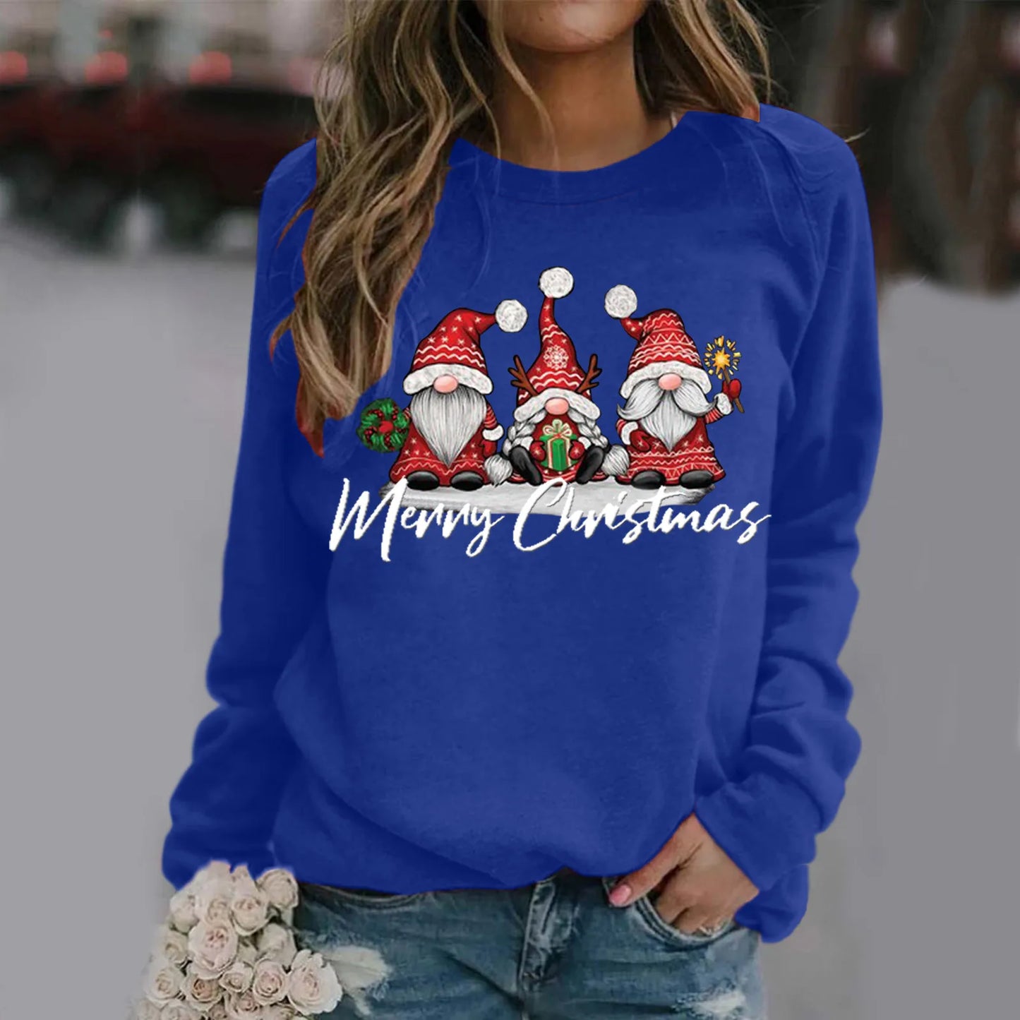 A woman holds flowers and wears a blue sweatshirt depicting three festive gnomes in red hats with Christmas accessories, and the words "Merry Christmas" in script.