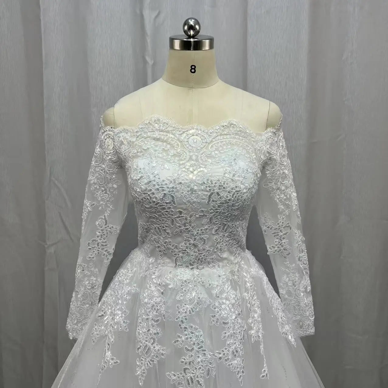 White lace wedding dress with intricate floral patterns on a mannequin. The dress features an off-shoulder design with long sleeves and a fitted bodice, set against a gray curtain backdrop.