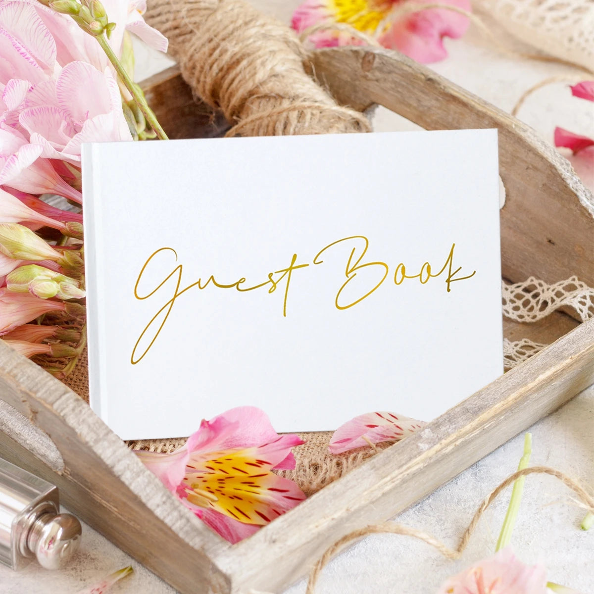 A white guest book with gold script sits in a wooden tray, surrounded by pink petals, lace, and twine, creating a rustic and elegant setting.