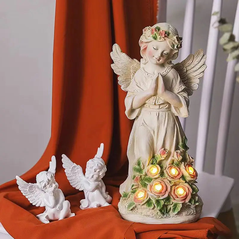 Ceramic angel statue with wings and flower crown, hands in prayer, surrounded by roses with glowing lights. Two smaller angel figurines sit nearby on an orange cloth.