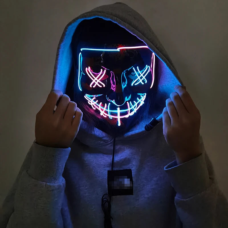 Person wearing a gray hoodie and a glowing neon mask with colorful X's over the eyes and a wide smile, holding the hood up, standing against a dark background.