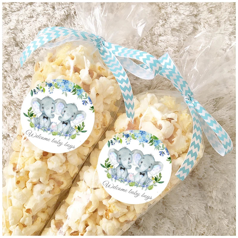 Clear gift bags filled with popcorn, tied with blue chevron ribbons, feature stickers of two elephants surrounded by flowers, reading "Welcome baby boys."