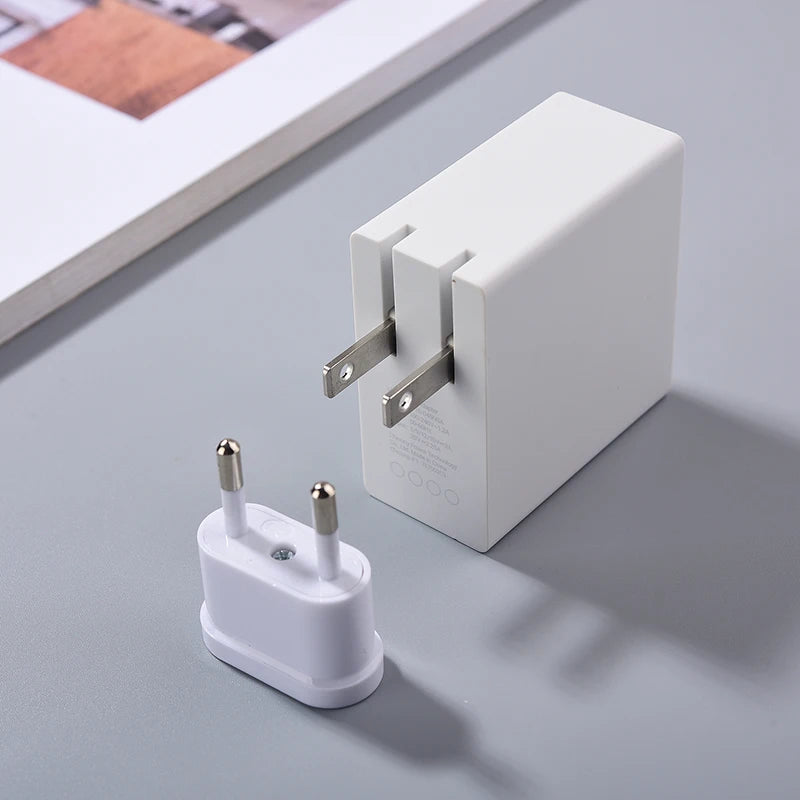 A white power adapter with dual flat prongs is next to a small white plug adapter with two round prongs, both on a gray surface near a book or magazine.