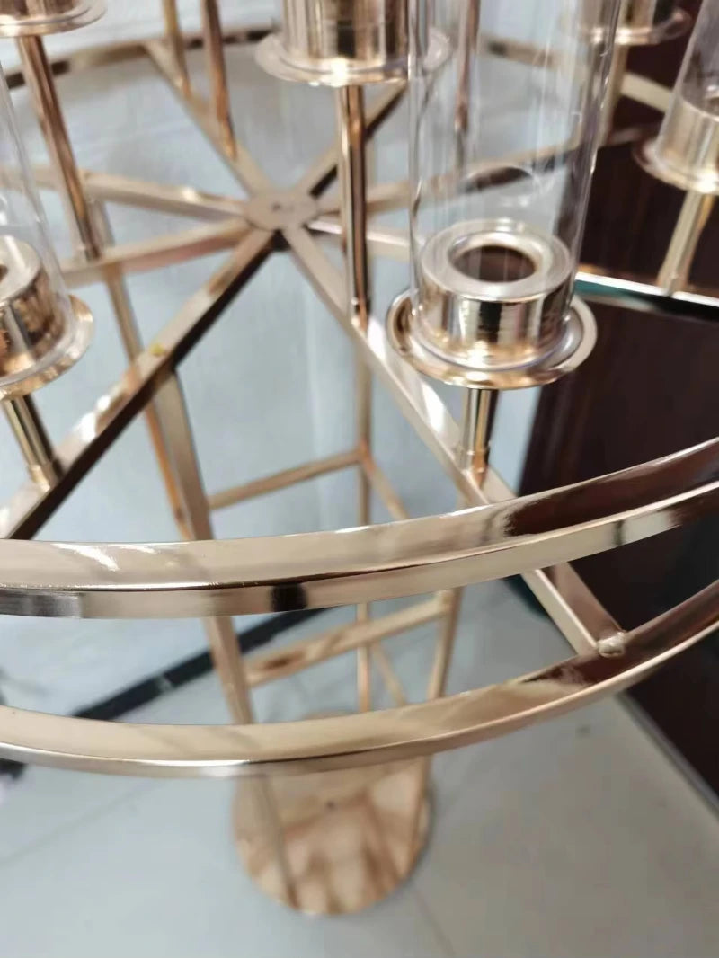 Close-up of a gold-colored, circular metal frame with six narrow, transparent cylindrical holders. The structure reflects light and is placed on a light-colored floor.