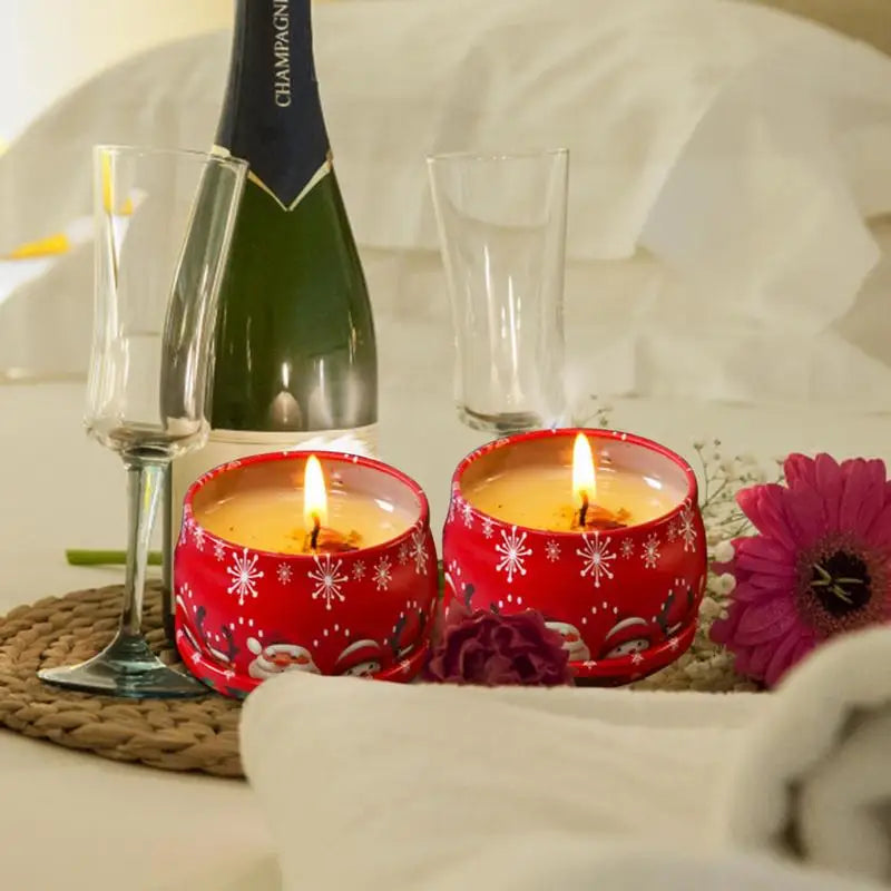 Cozy scene with two red candles, snowflake patterns, lit on a table near champagne, glasses, and pink flowers. Soft white cloth and gentle lighting create a romantic ambiance.
