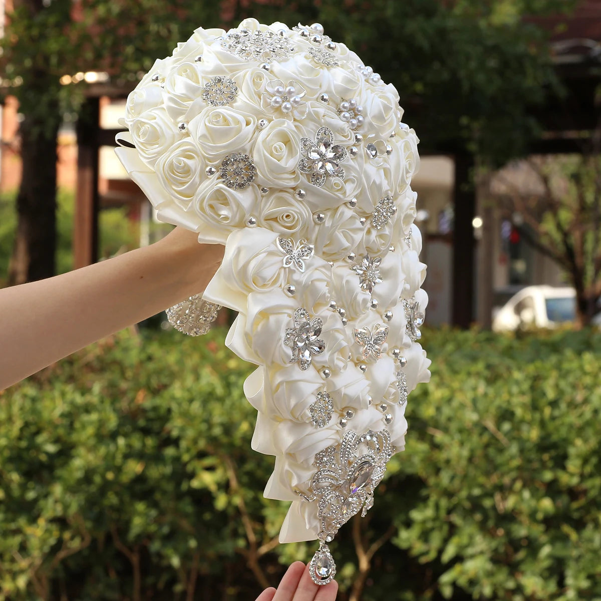 A hand holds a cascading bridal bouquet of white satin roses adorned with silver rhinestone brooches and pearls, set against a blurred garden background.
