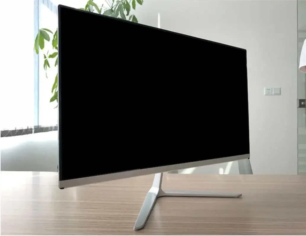 A sleek, modern flatscreen TV with a thin bezel and silver stand is positioned on a wooden table in a bright room with a large window and green leafy plant.