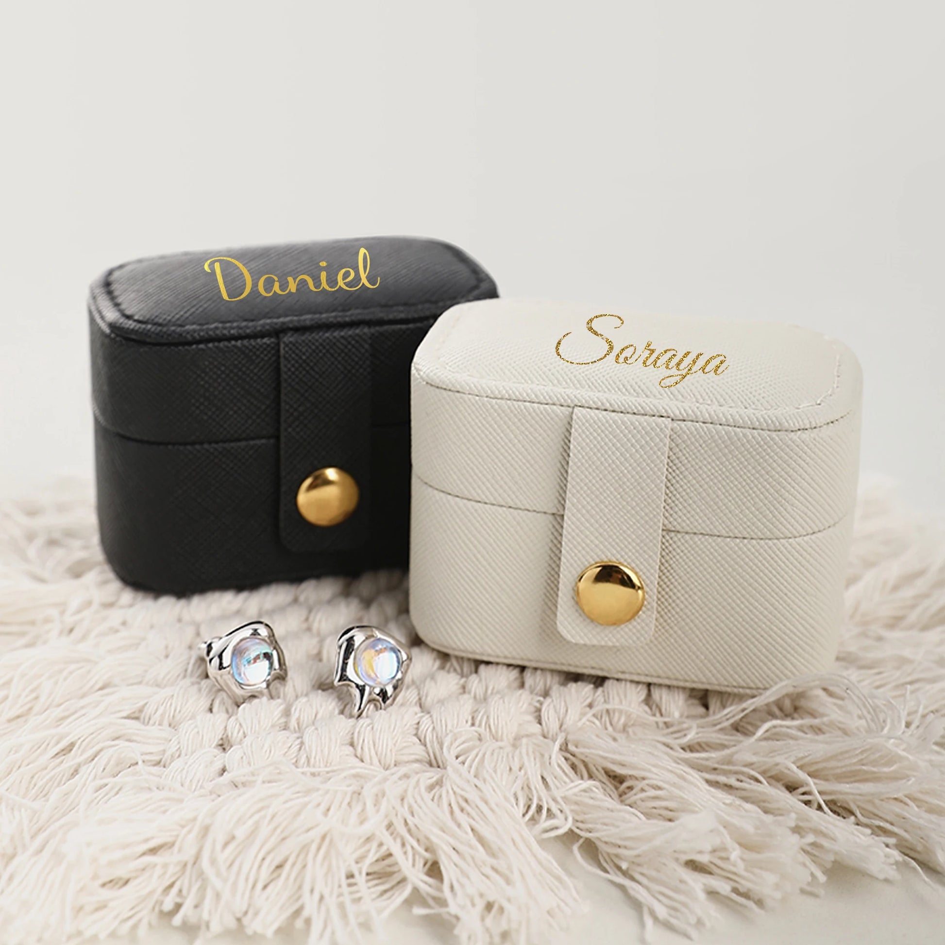 Two textured jewelry boxes, one black with "Daniel" and one white with "Soraya", sit on a woven cloth with a pair of silver earrings featuring iridescent stones in front.