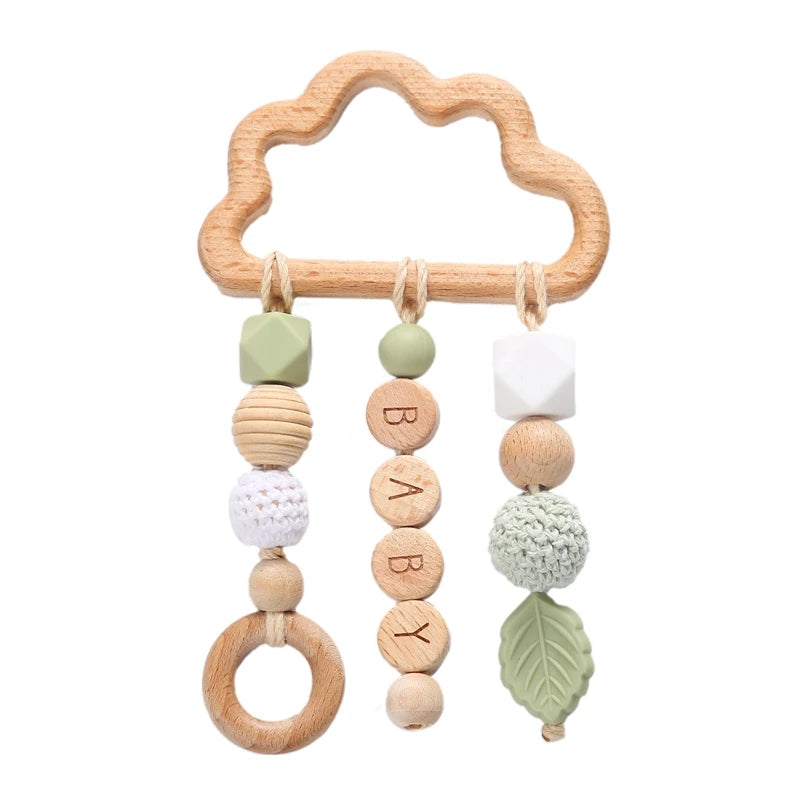 Wooden cloud-shaped baby toy with three hanging strands. Features green, white, and wood elements, including a ring, crocheted balls, leaf, and beads spelling "BABY".