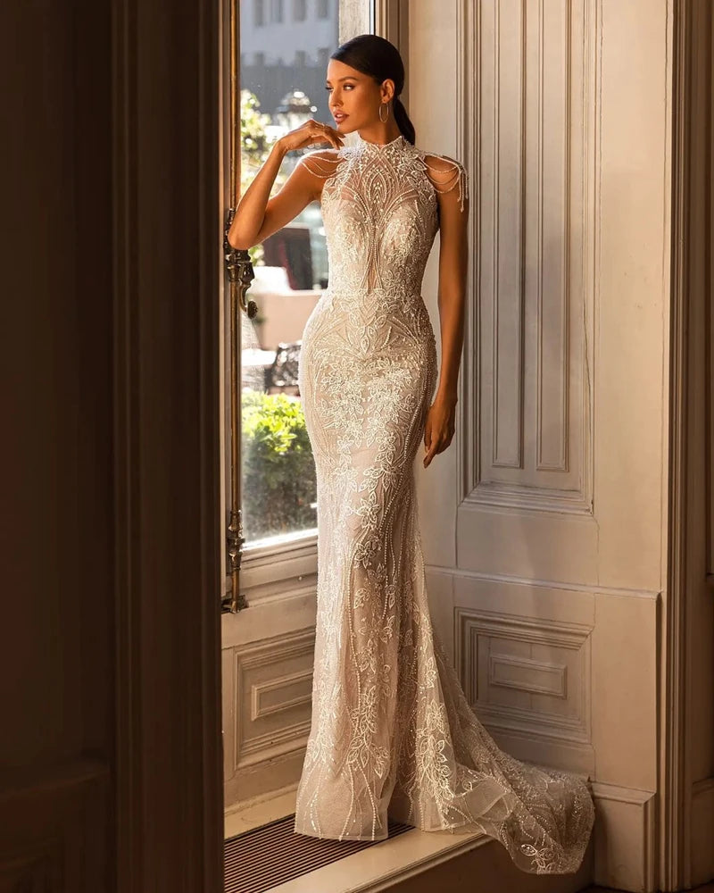 A woman stands gracefully in a sunlit room wearing a sleeveless, high-neck, intricately embroidered lace gown with a long train, looking out a tall window.