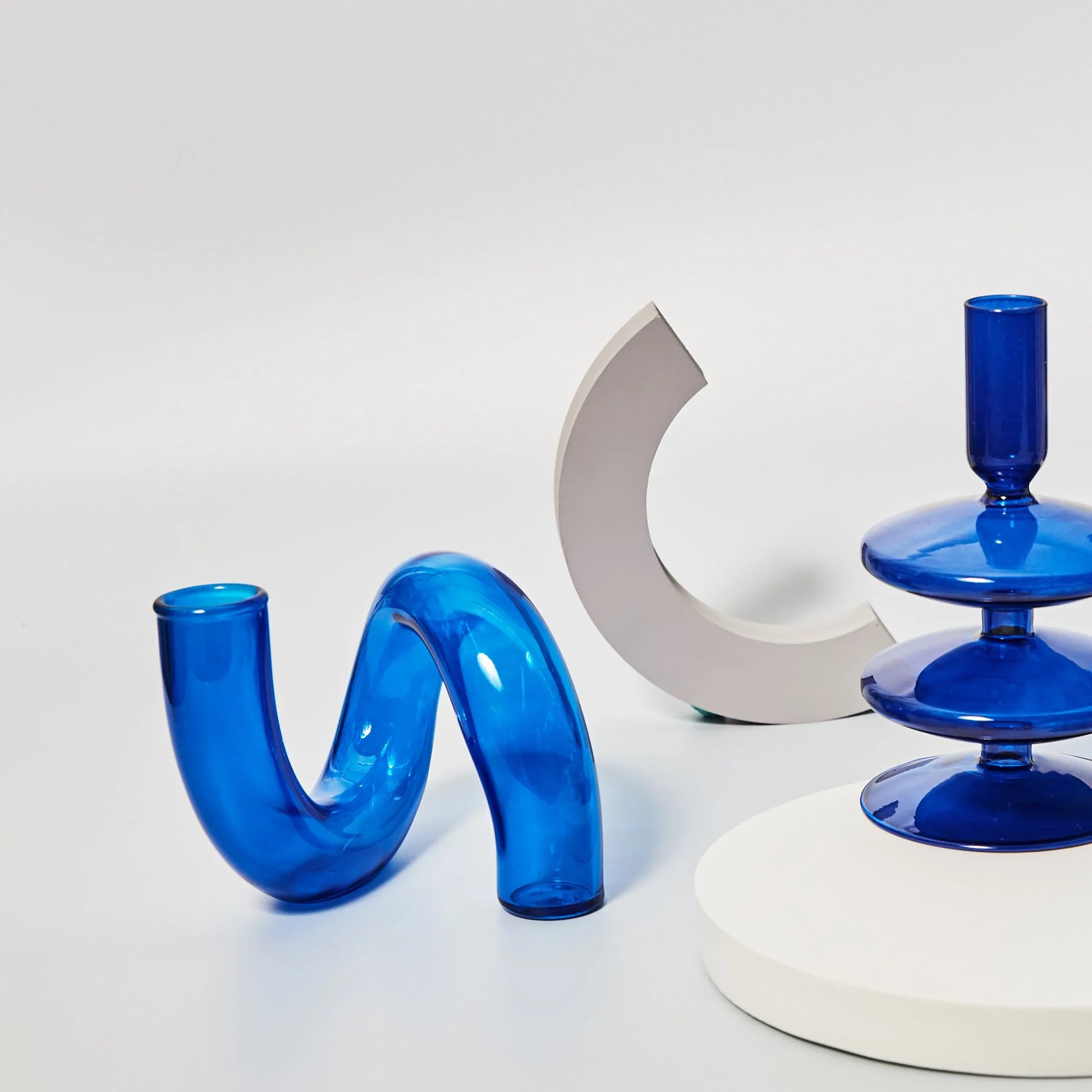 Curved translucent blue glass sculpture, a semicircular beige form, and a stacked circular blue glass piece on a white surface against a light background.