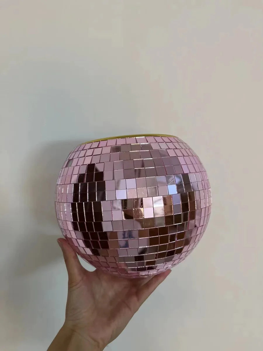 A hand holds up a pink mosaic mirror ball against a plain background, reflecting light in various shades of pink and gold.