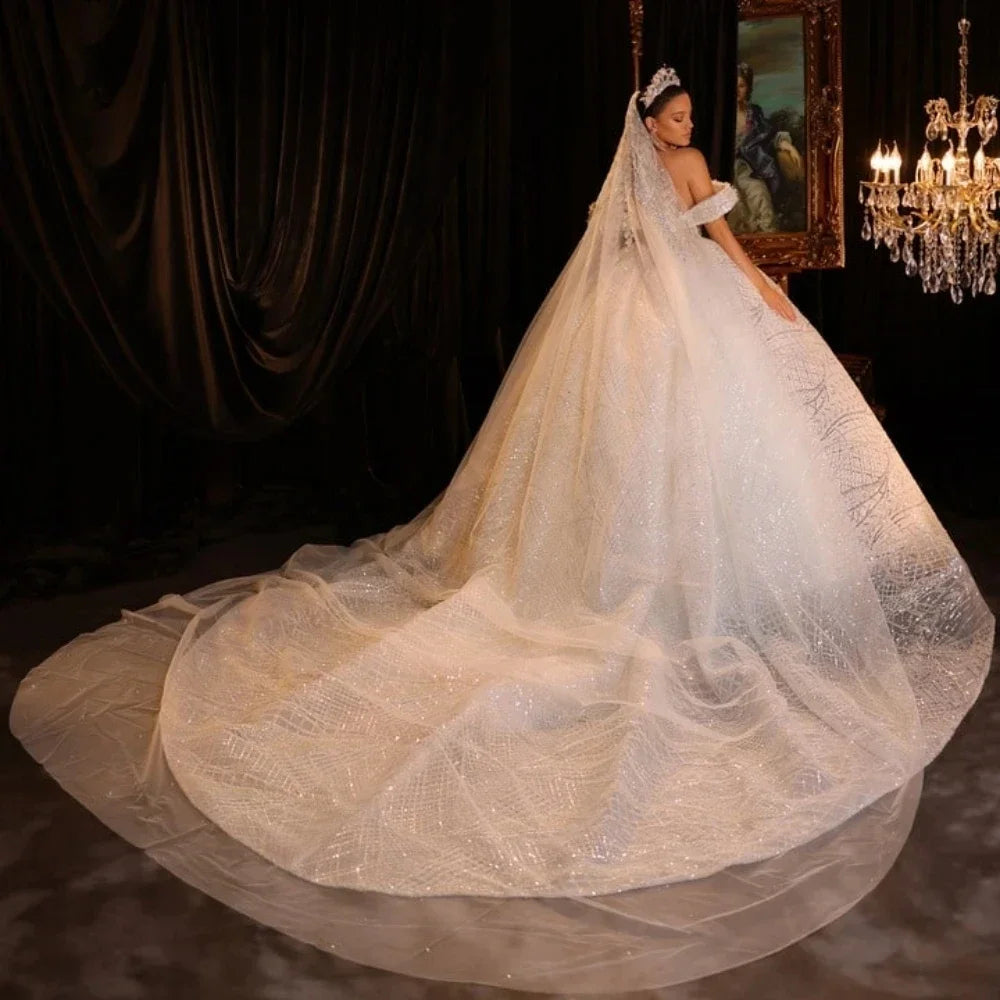 A bride in an elegant, off-shoulder wedding gown with a glittering, long train and veil stands beside a chandelier in a dimly lit room with dark drapes and a classic painting.