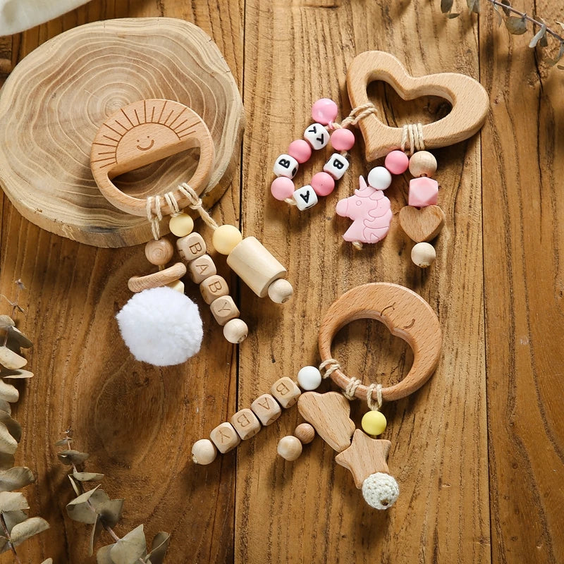 Wooden baby teethers on a wooden surface, featuring sun, heart, and moon shapes. Decorated with colorful beads, unicorn figure, and plush pom-poms. Blocks spell "BABY."