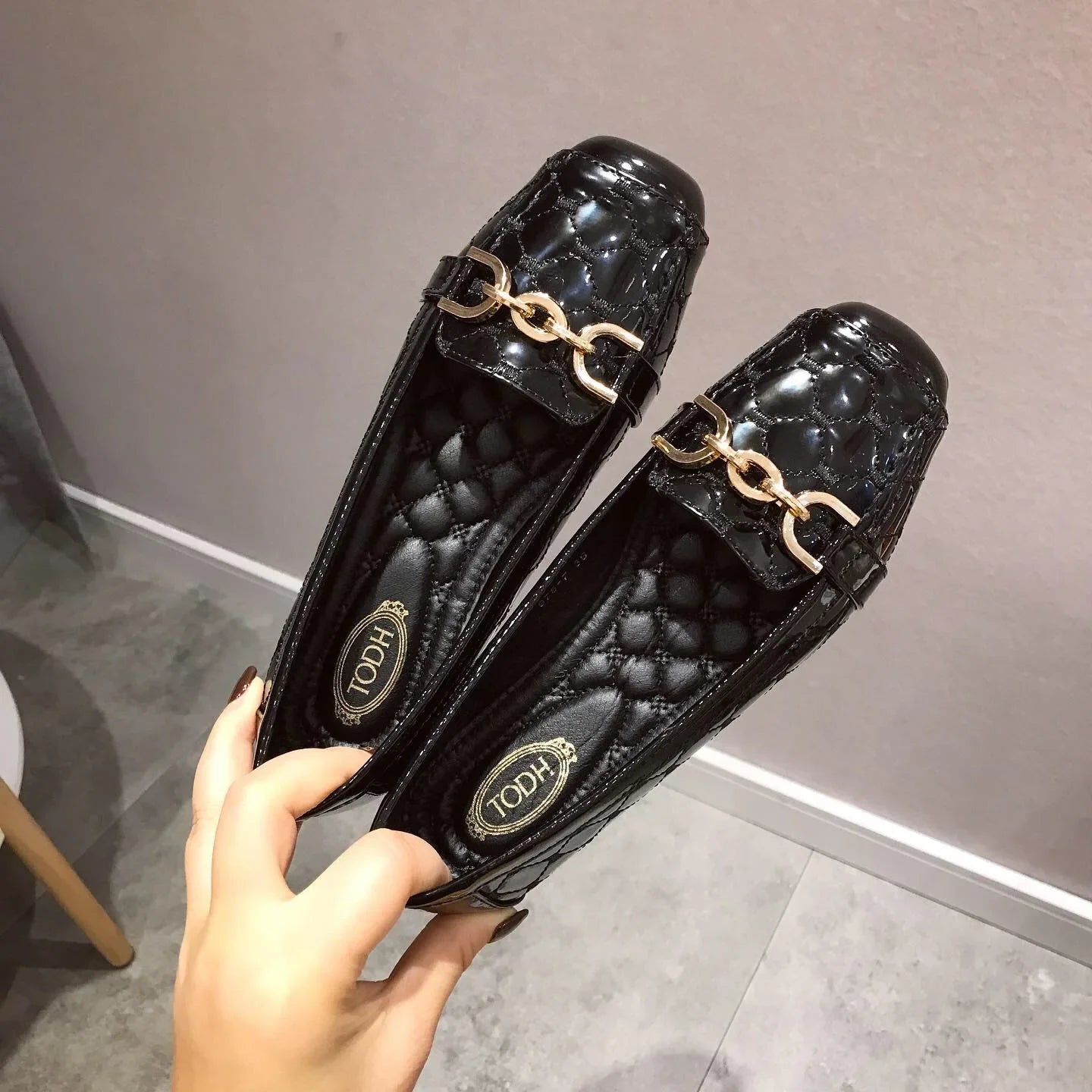 Pair of shiny black loafers with gold chain detail, quilted insole designs, held by a hand against a neutral background.