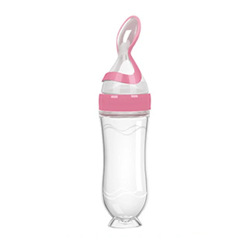 Clear squeezable baby food feeder with a pink cap and spoon-shaped silicone tip. The transparent body has wave patterns, and the base supports upright storage.
