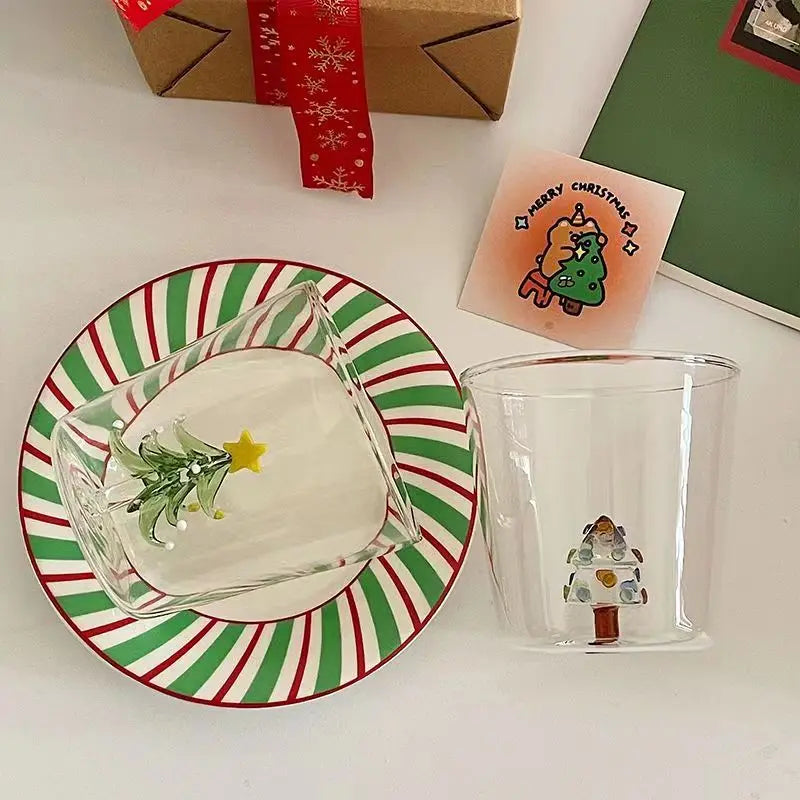 Two clear mugs with Christmas tree designs, a red and green striped plate, a green card, a Merry Christmas card with a cartoon tree, and a wrapped gift with red snowflake ribbon.