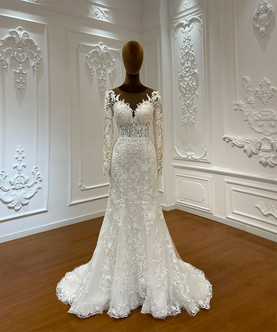 Elegant white lace bridal gown with long sleeves and intricate floral patterns, displayed on a mannequin in a tastefully decorated room with ornate white wall moldings.