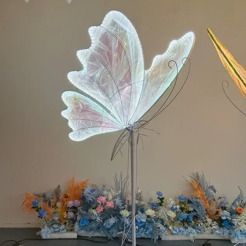 Illuminated butterfly wing sculpture with delicate wires stands amidst a colorful arrangement of artificial flowers, featuring shades of blue, pink, and orange.