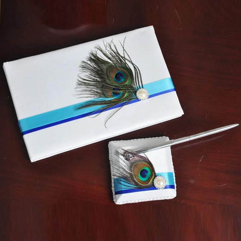 White guest book and pen holder on wood, adorned with blue ribbons, peacock feathers, and pearl accents. A silver pen rests on the holder.