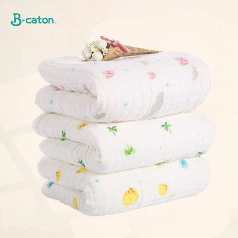 Three folded, white muslin baby blankets with colorful patterns of animals and plants, stacked neatly. A small bouquet of flowers rests on top. Logo in the corner.