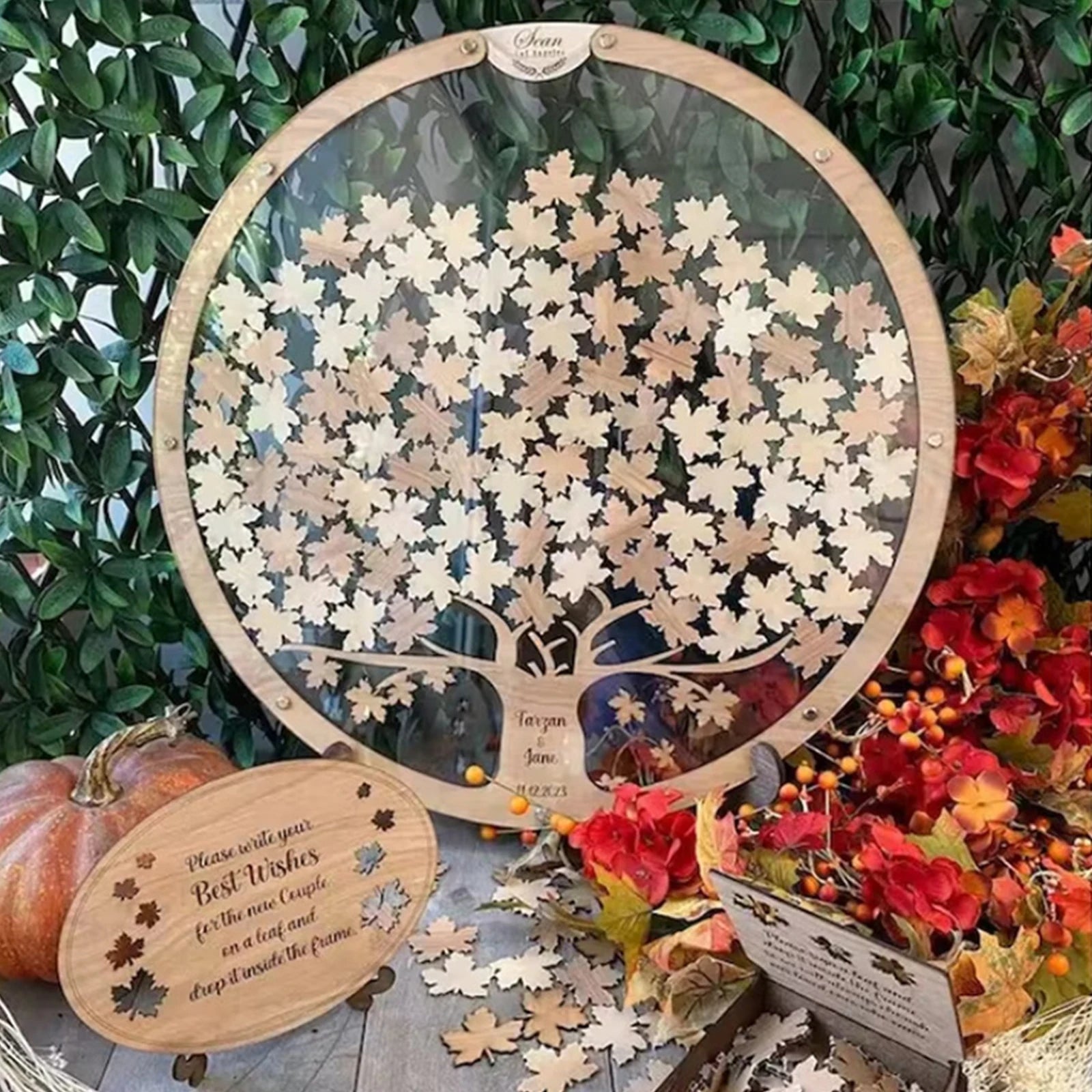 A round wooden frame holds a tree design with leaf-shaped notes. Nearby, a sign encourages best wishes for a couple. Autumn leaves and a pumpkin decorate the scene.