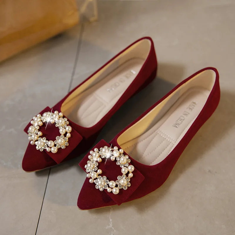 Elegant burgundy flats with pointed toes, adorned with pearl and rhinestone circular embellishments on the front. They rest on a gray tiled floor.