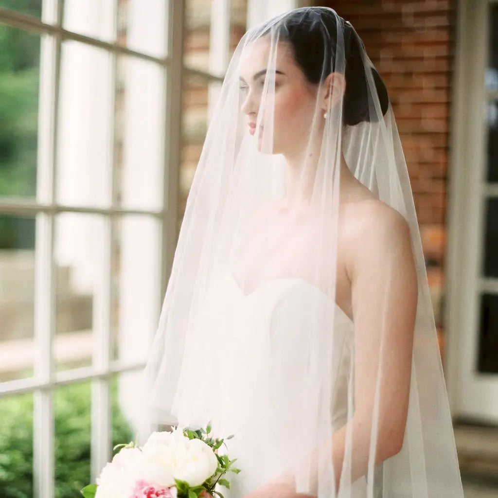 Bride in a strapless white gown and long veil, holding a bouquet of white and pink flowers, stands in front of a window with a brick wall background.