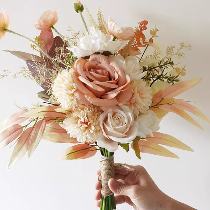 A hand holds a bouquet of artificial flowers including peach and white roses, cream-colored blooms, and delicate leaves, wrapped in twine against a light background.