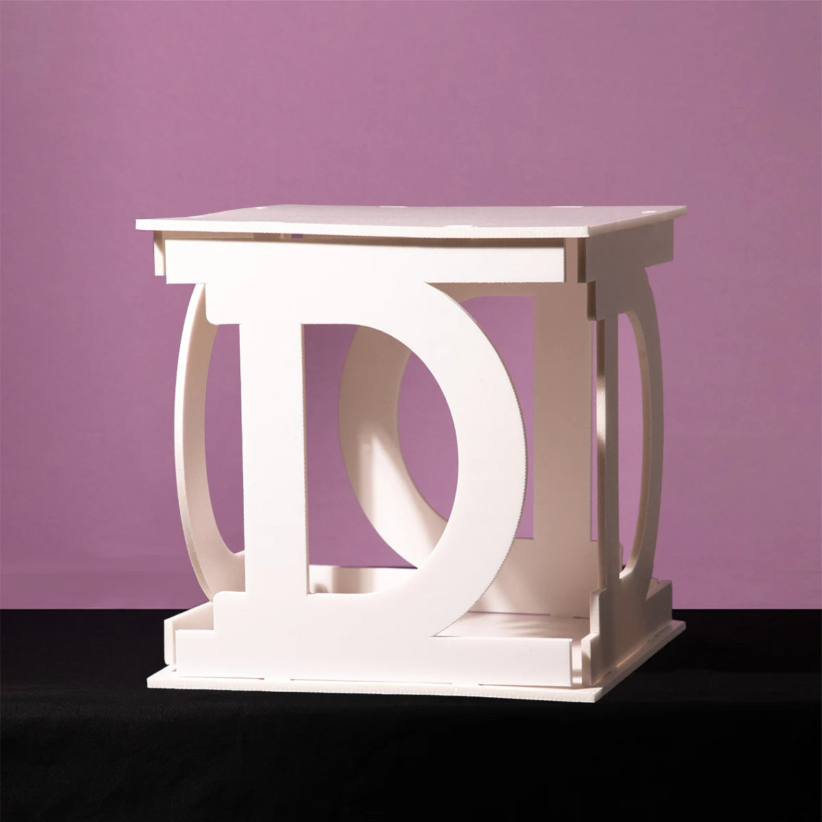 White geometric sculpture resembling interlocking "D" shapes against a purple background, placed on a dark surface.