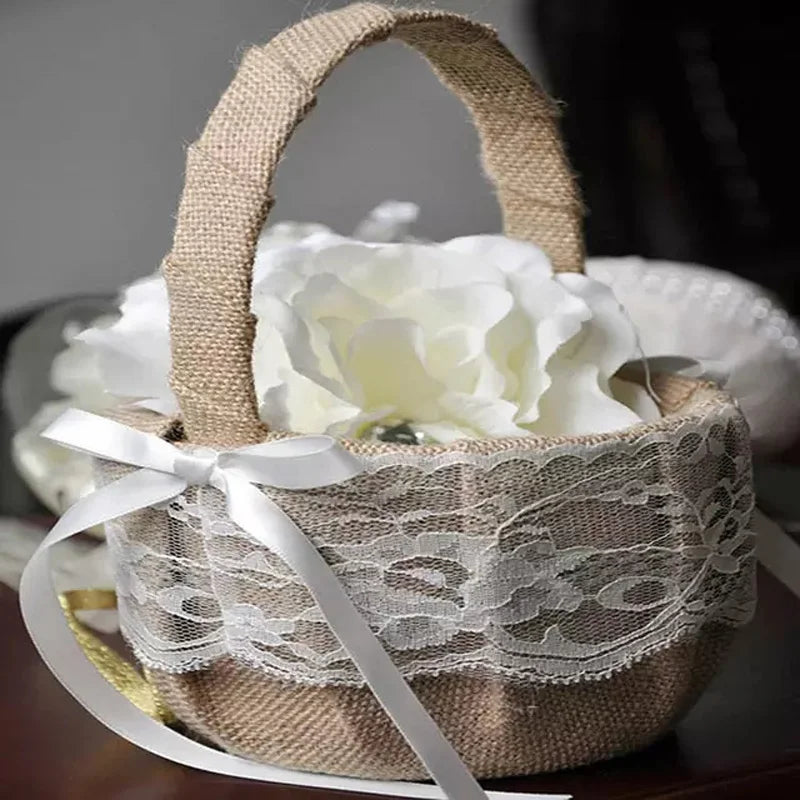 A small burlap basket adorned with lace and a white satin bow, containing a large white flower. The basket is elegantly displayed on a table.