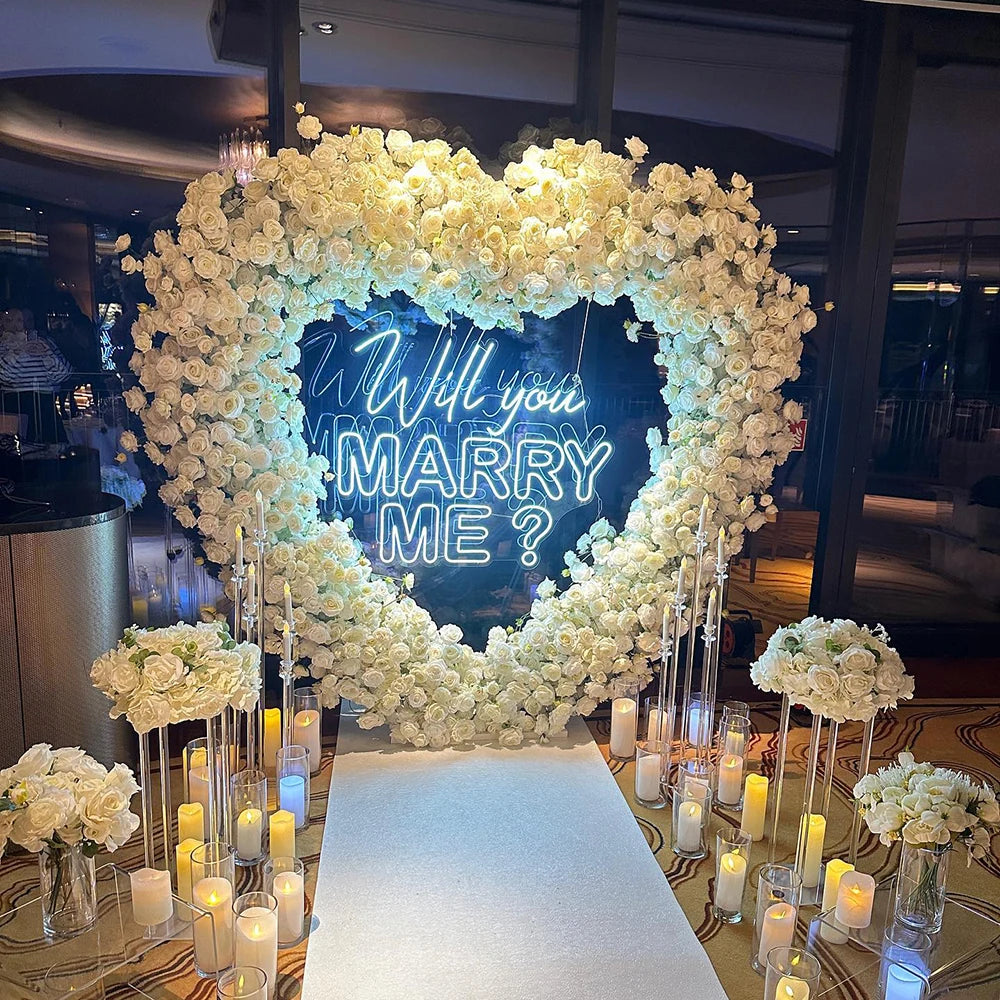 Heart-shaped floral arrangement with white roses surrounds a neon sign reading "Will you marry me?". Candles and bouquets line the aisle in an elegant indoor setting.