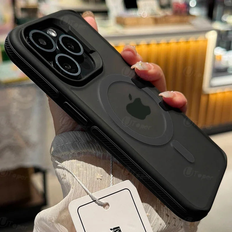 A hand holding a black iPhone with a rugged case featuring a visible Apple logo, triple camera setup, and a store ambiance blurred in the background.