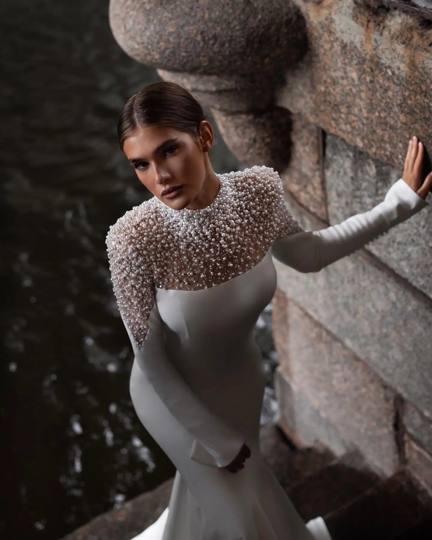 Woman in a long-sleeved, pearl-embellished gown stands by a stone wall, looking upward. Dark water is visible in the background, creating a dramatic contrast.