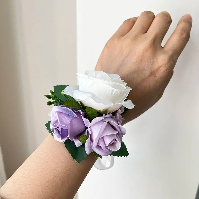 Wrist adorned with a floral corsage featuring a white rose and lavender roses, accented by green leaves. The background is a neutral, light-colored surface.