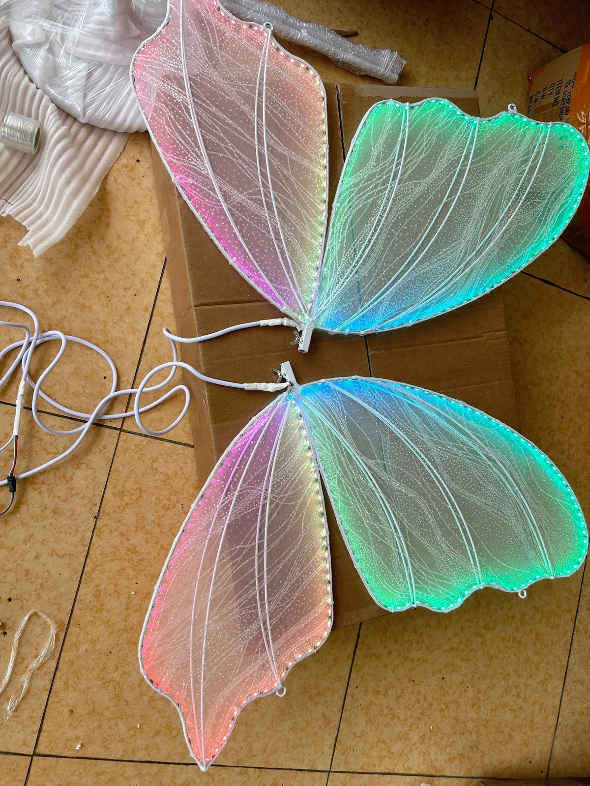 Colorful LED fairy wings with intricate patterns, emitting pink, blue, and green hues. Wings are placed on a cardboard box on a tiled floor, connected with white cords.