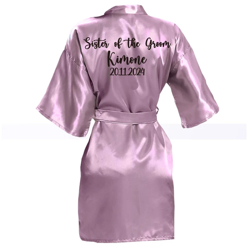 Lilac satin robe with "Sister of the Groom," "Kimone," and dates "2011.2024" written on the back in black script. The robe has wide sleeves and a matching belt.