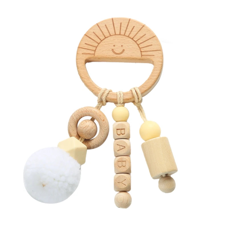 Wooden baby teether shaped like a smiling sun, featuring dangling elements: a wooden ring with a pom-pom, letter blocks spelling "BABY," and a cylindrical piece with beads.