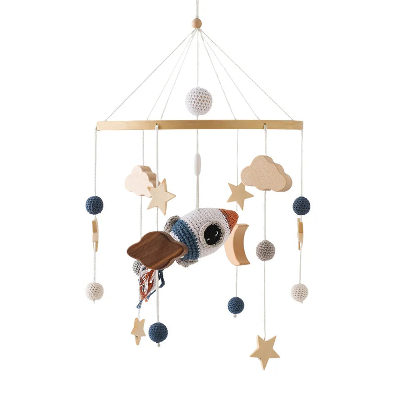 A baby mobile with a crocheted rocket, wooden stars, and clouds, hanging from a wooden frame. Accents include blue and beige crocheted balls, creating a whimsical space theme.