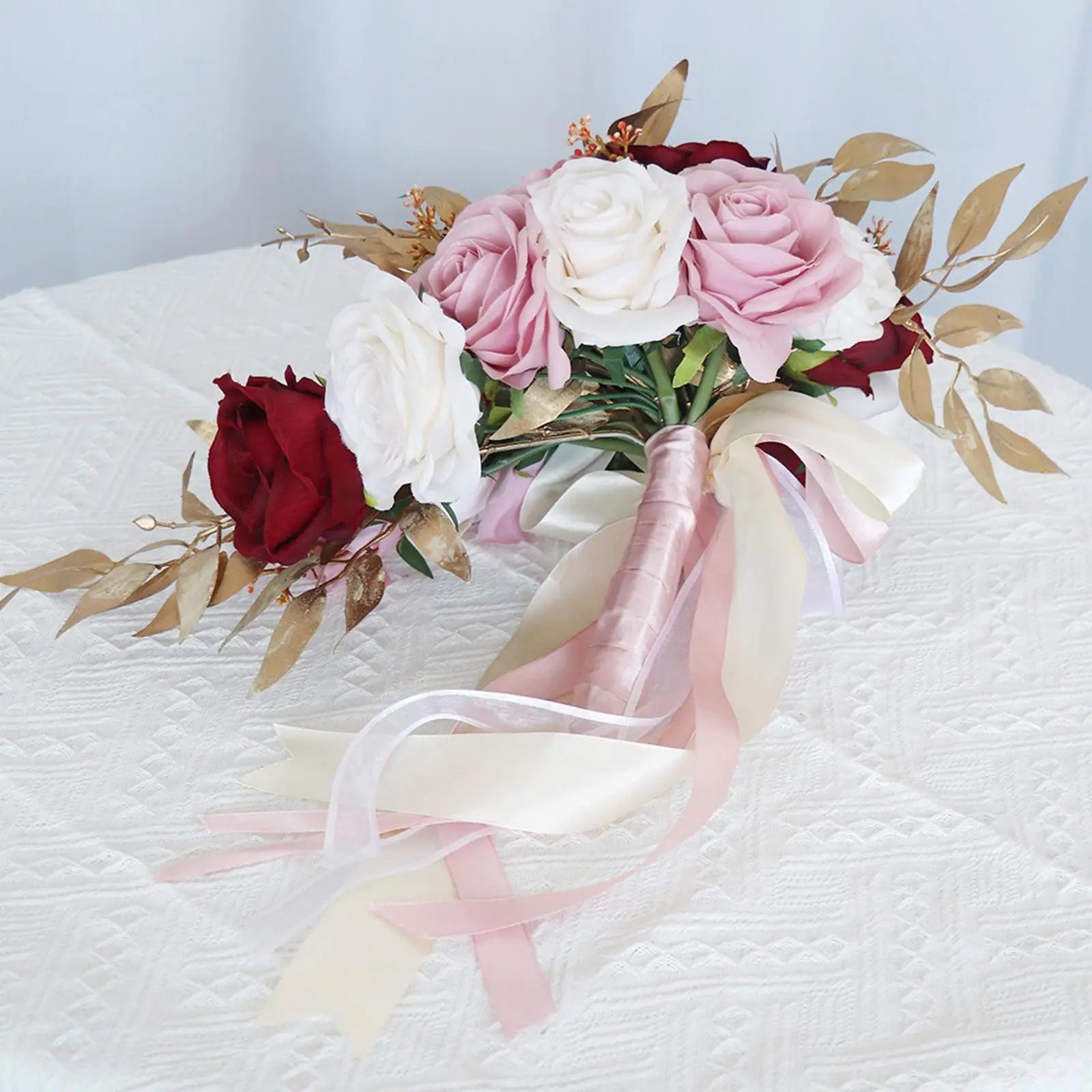 A bouquet of red, white, and pink roses with gold leaves and pink ribbons on a textured white tablecloth.