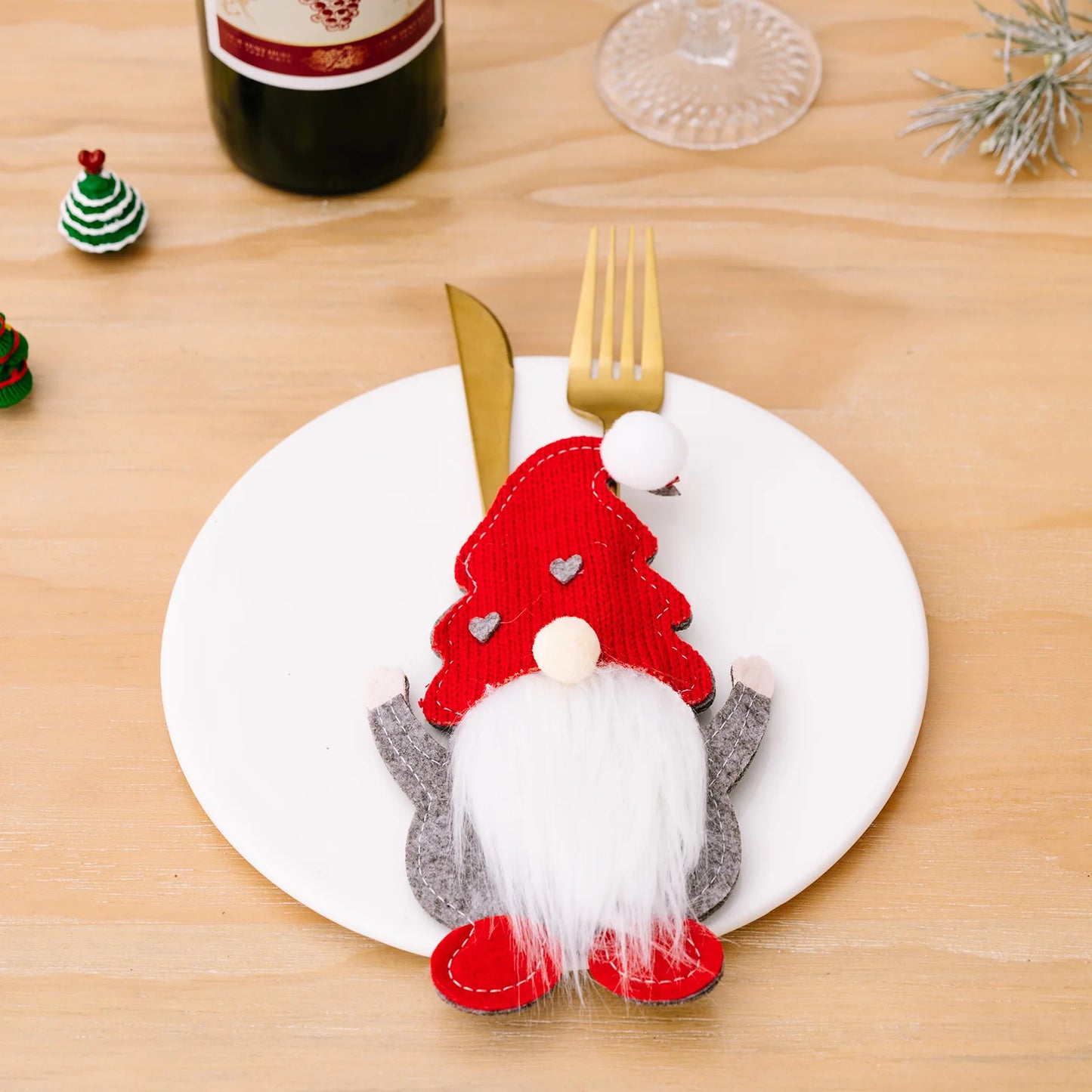 Festive table setting with a gnome-shaped felt cutlery holder on a white plate, holding a gold fork and knife. A wine bottle and mini Christmas trees decorate the wooden table.