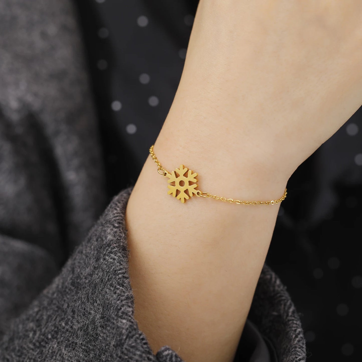 Gold snowflake charm bracelet on a wrist, with a gray sleeve visible, set against a blurred dark background with white dots.