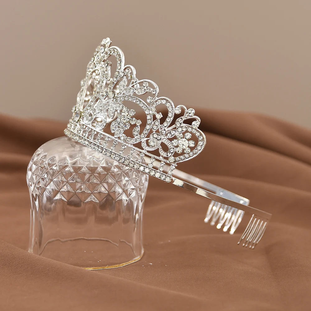 A sparkling silver tiara adorned with intricate patterns and clear gemstones, resting on a transparent glass with a brown fabric background.