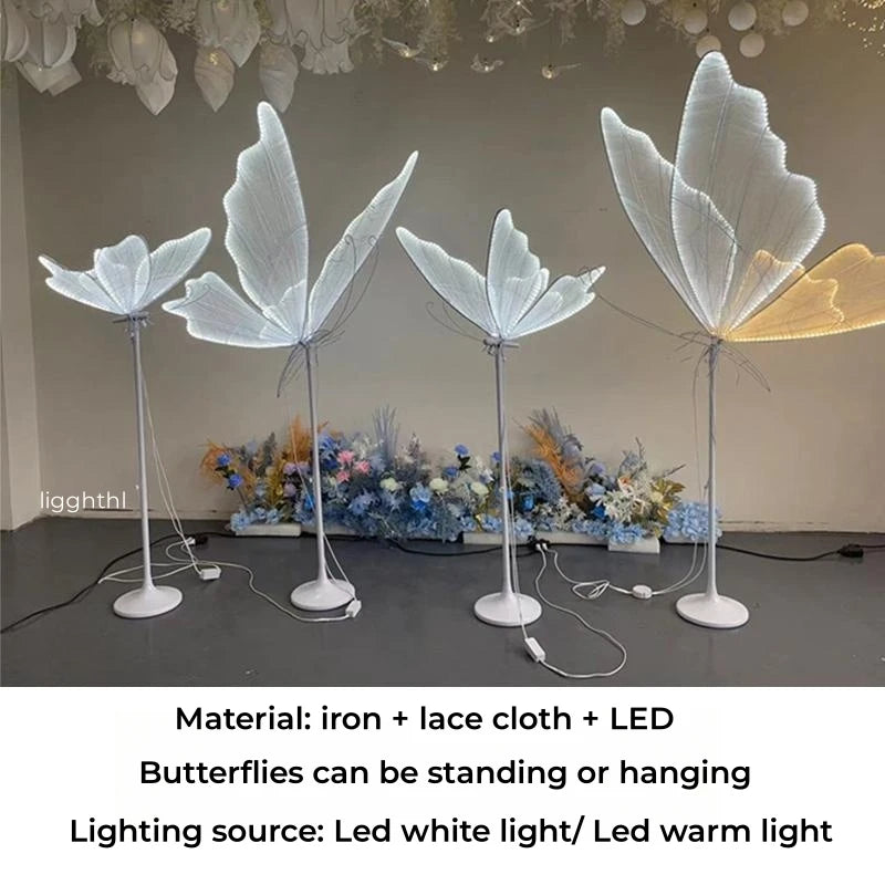 Four butterfly-shaped LED lamps made of iron and lace cloth stand on a floor, emitting soft white and warm light. Decorative flowers are placed behind them.