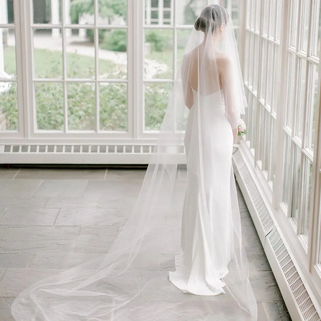 A bride in a sleek white gown and long veil stands by large windows, gazing outside. Light filters through, highlighting her elegant silhouette and creating a serene ambiance.