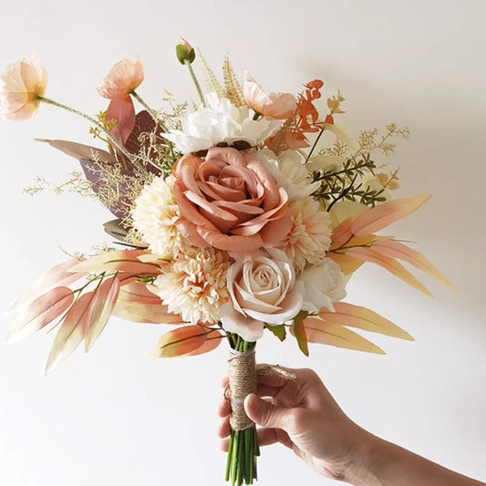 A hand holds a bouquet of flowers with peach and white roses, cream chrysanthemums, and green foliage. Wrapped stems add an earthy touch, set against a plain white background.