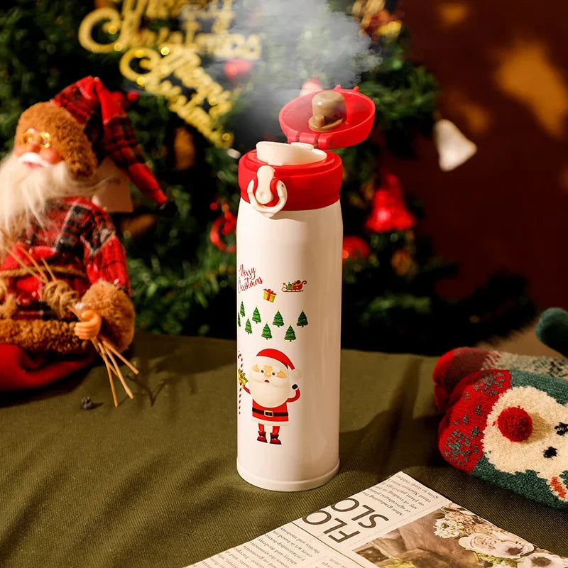 A festive white thermos with a Santa design on a green table, steaming under a Christmas tree. A Santa doll and a red knitted item are nearby, with holiday decorations in the background.