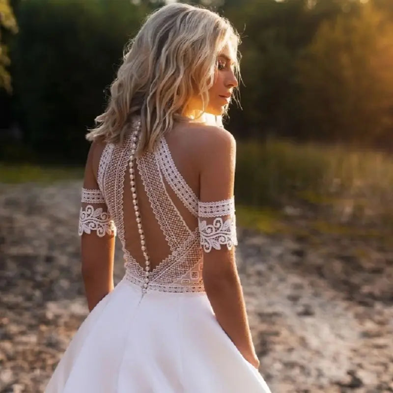 Bride in off-shoulder lace gown with intricate back design and pearl buttons stands outdoors in sunlight. Blond hair flows naturally, highlighting elegant dress details.