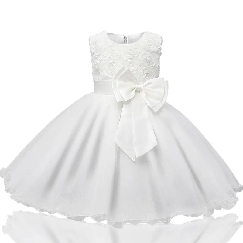 White sleeveless dress for a young child with a floral lace bodice and a large bow at the waist. Skirt is full and flared, creating an elegant, formal look.